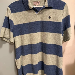 Randig frotté pikétröja från Morris - Säljer en snygg randig pikétröja från Morris i blått och beige. Tröjan har korta ärmar och en klassisk krage med en liten broderad detalj på bröstet. Perfekt för en avslappnad stil.