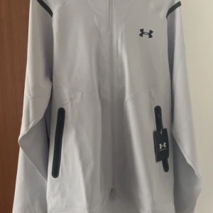 Grå jacka från Under Armour - Säljer en stilren grå jacka från Under Armour med dragkedja och justerbar huva. Jackan har svarta detaljer vid fickorna och logotypen på bröstet. Perfekt för en sportig look.helt fick som pressent då den var för stor splitter ny. Först till kvarn. Pris går att diskuteras  