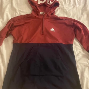 Röd och svart hoodie från Adidas - Säljer en snygg röd och mörk blå  hoodie från Adidas med halv dragkedja och stor ficka framtill. Perfekt för kyliga dagar med sin bekväma passform och stilrena design. Adidas-loggan pryder bröstet och huvan har en cool textdetalj. Passar både för träning och vardag.