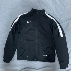 Svart vindjacka från Nike - Säljer en svart vindjacka från Nike med vit logga och vita ränder längs ärmarna. Jackan har en dragkedja framtill och är tillverkad i Dri-FIT material för optimal komfort. Perfekt för träning eller vardagsbruk. Storlek L men passar M bättre.