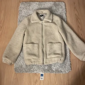Beige teddyjacka från Bubbleroom - Mysig beige teddyjacka från Bubbleroom med dragkedja framtill och två stora fickor. Perfekt för kyliga dagar när du vill hålla dig varm och stilren. Jackan har en klassisk krage och långa ärmar.