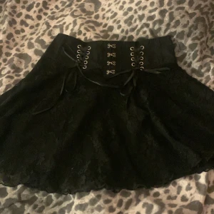 Svart kjol med snörning - Snygg svart kjol med snörning i midjan och spetsdetaljer. Perfekt för en cool och stilren look. Kjolen har en kort längd och en unik design som sticker ut.