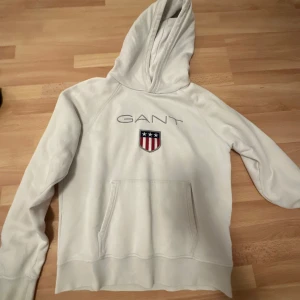 Vit hoodie från GANT - Säljer en stilren vit hoodie från GANT med en klassisk känguruficka och ett tryck med stjärnor och ränder på bröstet. Perfekt för en avslappnad look.