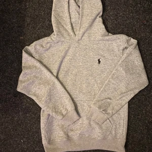 Grå hoodie från Ralph Lauren - Säljer en stilren grå hoodie från Ralph Lauren med klassisk logga på bröstet. Perfekt för en avslappnad look. Den har långa ärmar och en bekväm huva.