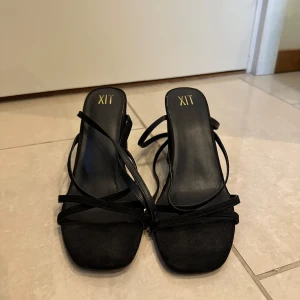 Svarta sandaler med klack från XIT - Eleganta svarta sandaler från XIT med hög klack och smala remmar. Perfekta för en stilren look med en touch av glamour. Remmarna ger en säker passform och den fyrkantiga tån ger en modern känsla.