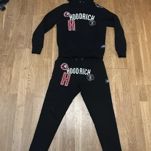 Svart hoodie från Hoodrich - Säljer en svart hoodie från Hoodrich med tryck i vitt och rött. Hoodien har en stor logotyp på bröstet och flera märken som ger en cool look. Perfekt för en avslappnad stil. (BYXORNA ÄR I STORLEK-M OCH TRÖJAN STORLEK-S