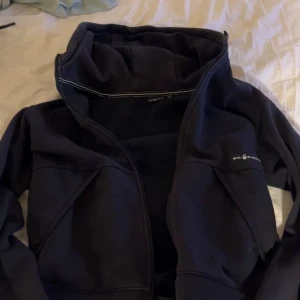 Svart hoodie från Sail racing - Svart hoodiejacka från Sail racing med dragkedja framtill och huva. Jackan har diskret logga på bröstet och är tillverkad i mjukt material, perfekt för lager-på-lager.