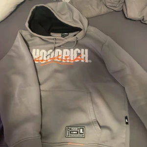 Grå hoodie från Hoodrich - Säljer en grå hoodie från Hoodrich i storlek L med vit och orange logga på bröstet. Tröjan har huva med dragsko, magficka och svart innerfoder i huvan. Perfekt för en avslappnad stil.
