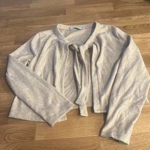 Beige kofta med knyt från 157 - Säljer en beige kofta från 157 i storlek XS. Koftan är croppad och har långa ärmar samt knyt framtill vid halsen. Perfekt för lager-på-lager och enkel att matcha med olika outfits.