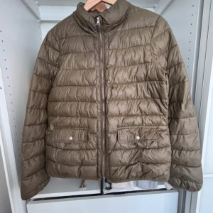 Olivgrön pufferjacka från Zara - Säljer en olivgrön pufferjacka från Zara med dragkedja framtill och två stora fickor med knappar. Jackan har en liten högre krage och quiltad design som ger en snygg och modern look. Har två små fläckar man knappt märker