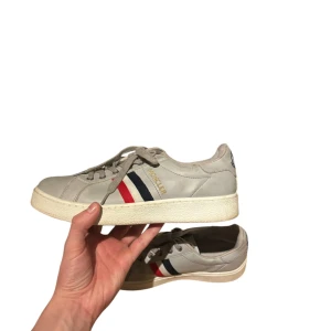 Gråa sneakers från Moncler - Säljer ett par gråa sneakers från Moncler med klassiska ränder i rött, vitt och blått på sidan. Skorna har snörning, rund tå och vit platt sula. Moncler-logga på hälen. Perfekta för dig som gillar stilrena och sportiga skor. Skorna är storlek 42. 