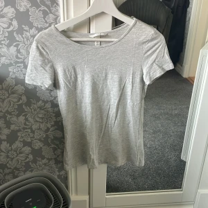 Ljusgrå t-shirt från H&M - En basic ljusgrå t-shirt från H&M med rund halsringning och korta ärmar. Perfekt till jeans eller shorts och enkel att matcha med andra plagg. Tillverkad i mjuk bomull för en bekväm känsla.