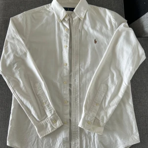 Vit skjorta från Ralph Lauren - Klassisk vit skjorta från Ralph Lauren med broderad logga på bröstet. Skjortan har lång ärm, knappar framtill och är tillverkad i bomull. Perfekt för en stilren och tidlös look.