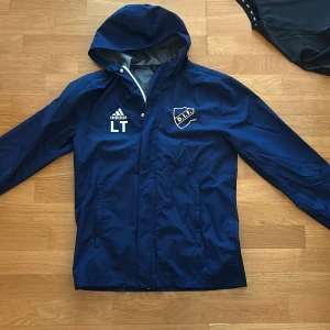 Blå regnjacka från Adidas x DIF - Säljer en blå vindjacka med huva från Adidas i samarbete med Djurgårdens IF. Jackan har vitt tryck med klubbmärke, där det har gått av lite från märket men går att fixas med lite lim. och en Adidas-logga på bröstet samt Stadium-tryck på ryggen. Perfekt för fotbollsträning eller som supporterplagg.