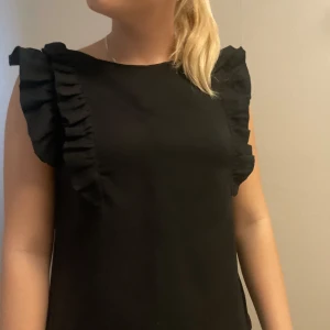 Svart topp med volangärmar - Säljer en svart topp med volangdetaljer längs ärmarna. Toppen har en rak passform och är ärmlös, vilket gör den enkel att styla till olika outfits. Perfekt för dig som gillar stilrena plagg med en twist.