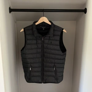 Ralph Lauren väst  - Säljer en grå/svart dunväst från Polo Ralph Lauren med dragkedja framtill och två fickor med dragkedja. Västen har en klassisk, quiltad design och hög krage. Perfekt för lager-på-lager-stil. Skicka privat vid minsta fundering eller fråga!