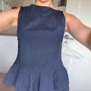 Mörkblå randig topp  med peplum - Säljer en mörkblå topp med smala vita ränder och peplumdetalj i midjan. Ärmlös modell med rund halsringning och figurnära passform upptill. Perfekt för dig som gillar klassisk och stilren look.