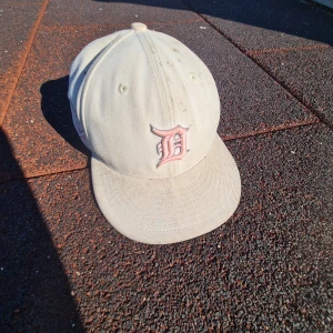 Beige keps från New Era 59FIFTY - Säljer en beige keps från New Era 59FIFTY med broderad bokstav på framsidan och ett broderat motiv på sidan. Kepsen har klassisk form och platt skärm. Passar dig som gillar stilrena accessoarer med unika detaljer.