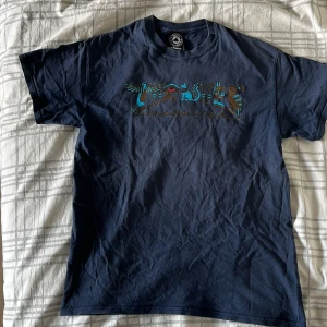 Mörkblå Thrasher t-shirt - Säljer en mörkblå t-shirt från Thrasher med färgglad logga och grafik på bröstet. Klassisk passform och rund halsringning. Perfekt för dig som gillar streetwear och skatekultur. Hål i högra armhålan.