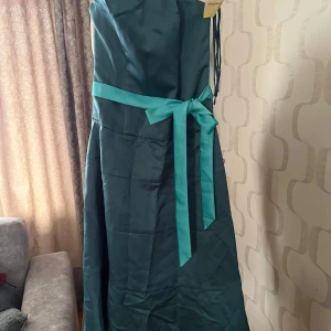 Mörkgrön axelbandslös långklänning med midjeband - Elegant mörkgrön axelbandslös långklänning med matchande band i midjan som knyts till rosett. Klänningen har en slät, glansig yta och en klassisk siluett som passar perfekt till festliga tillfällen.