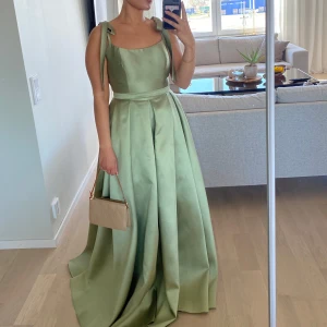 Ljusgrön långklänning med rosettband - Elegant ljusgrön långklänning med breda axelband som knyts i rosetter. Klänningen har markerad midja med bälte och vacker, voluminös kjol. Perfekt för bal eller fest. Tillverkad i glansigt satinliknande material.