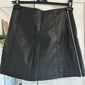 Svart skinnkjol med dragkedja från H&M Divided - Säljer en svart kort skinnkjol från H&M Divided i storlek 36. Kjolen har en snygg sned dragkedja framtill som detalj och är tillverkad i ett slätt, syntetiskt skinnmaterial. Perfekt för dig som gillar en stilren och edgy look.