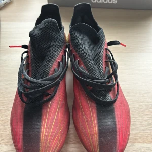 Adidas X Speedflow fotbollsskor i rött och guld - Säljer ett par Adidas X Speedflow fotbollsskor i rött med svarta och guldiga detaljer. Skorna har snörning, låg profil och guldiga dobbar för bästa grepp på planen. Perfekta för dig som vill sticka ut med stil och prestanda på fotbollsplanen.
