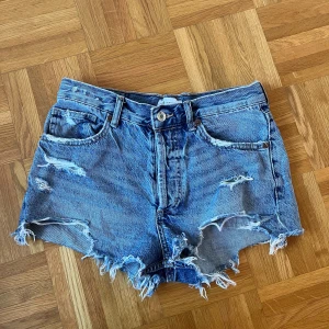 Blå jeansshorts från Zara - Säljer ett par blå jeansshorts från Zara i storlek 36 (S). Shortsen har fransiga benslut och slitna detaljer framtill och baktill. Klassisk femficksmodell.