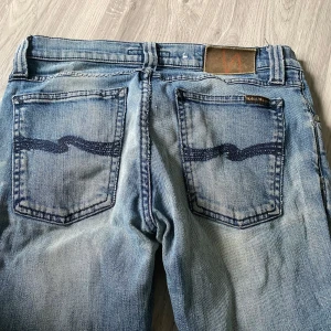 Blå jeans från Nudie Jeans - Säljer ett par klassiska blå jeans från Nudie Jeans med tydliga slitningar och signaturbroderi på bakfickorna. Modellen har raka ben och normal passform. Perfekta för dig som gillar en avslappnad stil. Hål vid knäna men det är en del av designen. Priset är inte hugget i sten