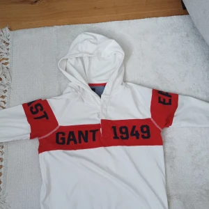 Vit och röd hoodie från GANT - Säljer en snygg vit hoodie från GANT med röda paneler och svart text. Tryck med 'GANT 1949' framtill och 'CONNECTICUT' på ryggen. Hoodien har långa ärmar och en klassisk huva. På sidan av armarna står det 'EAST' och på den andra 'COAST'Perfekt för dig som gillar sportig stil.