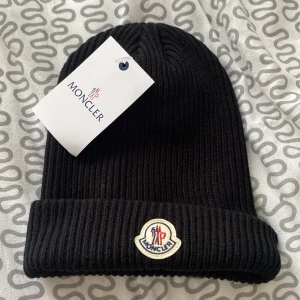 Svart ribbad mössa från Moncler - Svart ribbstickad mössa från Moncler med uppvikt kant och klassisk Moncler-logga framtill. Perfekt accessoar för kyliga dagar och enkel att matcha med olika outfits.