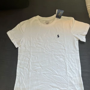 Vit t-shirt från Polo Ralph Lauren - Klassisk vit t-shirt från Polo Ralph Lauren med rund hals och korta ärmar. Liten broderad logga i marinblått på bröstet. Perfekt basplagg i mjuk bomull.