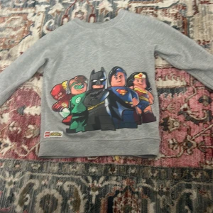 Grå långärmad tröja med LEGO superhjältar - Grå långärmad tröja med tryck av LEGO-versioner av superhjältar som Batman, Superman, Wonder Woman, Green Lantern och Flash på framsidan. Perfekt för dig som gillar superhjältar och LEGO! För små barn storlek 134-140 