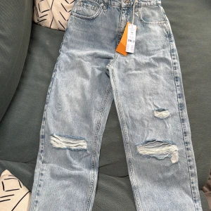 Straight 90s jeans med slitningar - Ljusa blå jeans i 90-talsstil med raka ben och hög midja. Byxorna har slitna detaljer på båda benen framtill och klassiska fem fickor. Perfekta för en avslappnad och trendig look. Jeansen går inte längre att köpa. Dom är aldrig anvönda💞