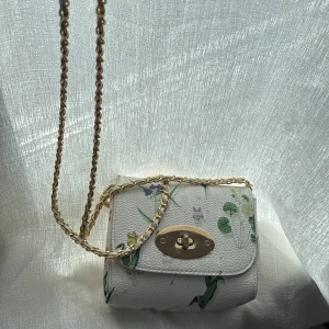 Vit axelväska med blommigt mönster och guldkedja - Superfin liten axelväska i vitt med blommigt mönster och guldfärgad kedja. Väskan har ett snyggt vridlås i guld på framsidan och är perfekt för att lyfta vilken outfit som helst. Den är kompakt och smidig att bära med sig.