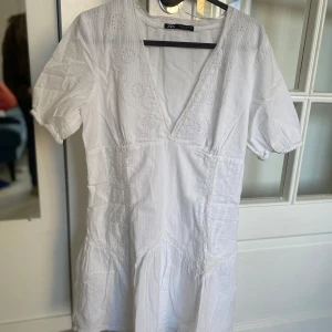 Vit v-ringad kortklänning  - Superfin vit klänning från Zara med v-ringning och kort ärm. Klänningen har subtila broderade detaljer upptill och en lätt volang nedtill. Perfekt för sommarens alla tillfällen!