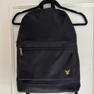 Svart ryggsäck från Lyle & Scott - En stilren svart ryggsäck från Lyle & Scott med klassisk design. Väskan har ett stort huvudfack, en ytterficka med dragkedja och justerbara axelremmar. Liten gul logotyp med örn på framsidan. Perfekt för skola eller vardag.
