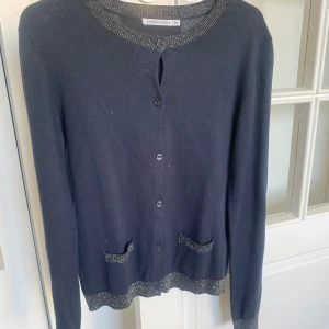 Mörkblå kofta med glittriga detaljer - Säljer en mörkblå kofta från Monoprix med glittriga silverdetaljer vid krage, fickor och nederkant. Koftan har långa ärmar och knäpps framtill med knappar. Perfekt att slänga över en topp för en snygg look.