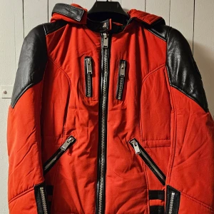 Röd och svart dam dun jacka från Horspist - Säljer en riktigt snygg röd dam/ barn jacka från Horspist med svarta detaljer i skinnimitation på axlar, armbågar och rygg. Jackan har flera dragkedjor framtill, huva och coola spänndetaljer bak. Perfekt för dig som vill sticka ut. Jackan är mycket mindre I storleken/ motsvarar mindre S. Finns även stor päls till luvan om så önskas. Luvan är avtagbar.
