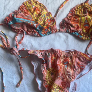 Mönstrad bikini med knyt - Säljer en färgglad bikini med paisley-inspirerat mönster i rosa, gult och blått. Både överdel och underdel har knytband i sidorna och vid nacken. Helt oanvänt! Storlek S!