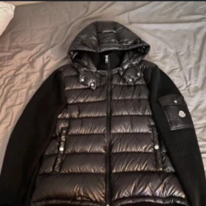 Moncler cardigan  - Säljer en svart pufferjacka från Moncler med stickade ärmar och huva. Jackan har dragkedja framtill, fickor med knappar och Moncler-logga på ärmen. Perfekt för kyliga dagar och riktigt snygg streetstil. Jackan har varit använd men är i okej skick 