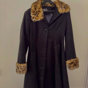 Svart kappa med leopardfuskpäls från Hell Bunny - Säljer en svart kappa från Hell Bunny med bred krage och ärmslut i leopardmönstrad fuskpäls. Kappan har knäppning framtill och markerad midja med knappdetalj bak. Perfekt statement-plagg för dig som gillar retrostil!
