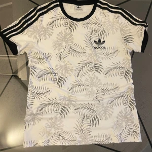 Vit Adidas t-shirt med bladprint - Säljer en fräsch vit Adidas t-shirt med svarta detaljer och grått bladmönster. Klassiska svarta ränder på axlarna och Adidas-logga på bröstet. Perfekt för dig som gillar sportig stil och snygga prints.