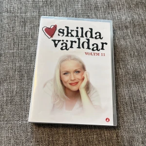 Skilda världar Volym 11 - DVD-box med Skilda världar Volym 11. Omslaget är vitt med en bild på en kvinna med ljust hår och röd läppstift. Serienamnet står i svart text med ett rött hjärta. Perfekt för dig som gillar svenska dramaserier.