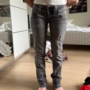 Grå slitna jeans med raka ben - Säljer ett par grå jeans med raka ben och slitningar på låret och knät. Klassisk femficksmodell med låg midja och dragkedja. Perfekta för en avslappnad stil.