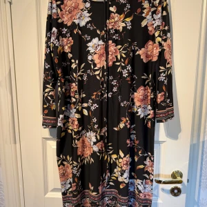 Svart blommig långklänning - Svart långklänning med stora blommiga mönster i rosa, beige och vitt. Klänningen har långa ärmar och dekorativa bårder vid ärmslut och nederkant. Perfekt för dig som gillar romantiska och färgstarka plagg.