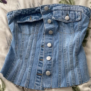 Jeans korsettopp med knappar - Säljer en snygg korsettopp i ljusblå denim med silvriga knappar framtill och två dekorativa fickor upptill. Toppen har smock baktill för bättre passform och råa kanter nedtill för en trendig look. Aldrig använd