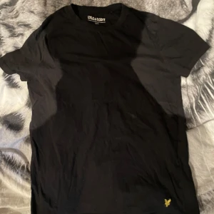 Svart t-shirt från Lyle & Scott. Nypris 499 säljer för 110 - Säljer en klassisk svart t-shirt från Lyle & Scott med deras ikoniska gula logga broderad längst ner på framsidan. T-shirten har rund hals och korta ärmar, perfekt för en clean och enkel stil.