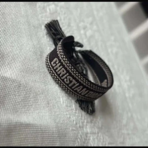 Svart armband från Dior med tofsar - Säljer ett svart tygarmband från Dior med texten 'CHRISTIAN DIOR J'ADIOR' broderad i vitt. Armbandet har dekorativa tofsar i ändarna och justerbar knytning. Perfekt accessoar för att lyfta din outfit.