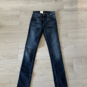 Mörkblå jeans High Kai från Nudie Jeans - Säljer ett helt par nya mörkblå jeans från Nudie Jeans, modellen High Kai. Jeansen har klassisk femficksdesign, raka ben och snygga detaljer med broderad bakficka och läderpatch i midjan. Perfekta för dig som gillar stilrena och tidlösa jeans.